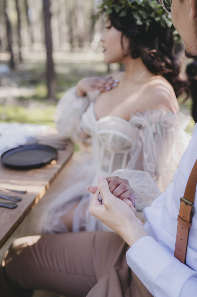 A boho bride and groom holding hands at their table in the forest.
