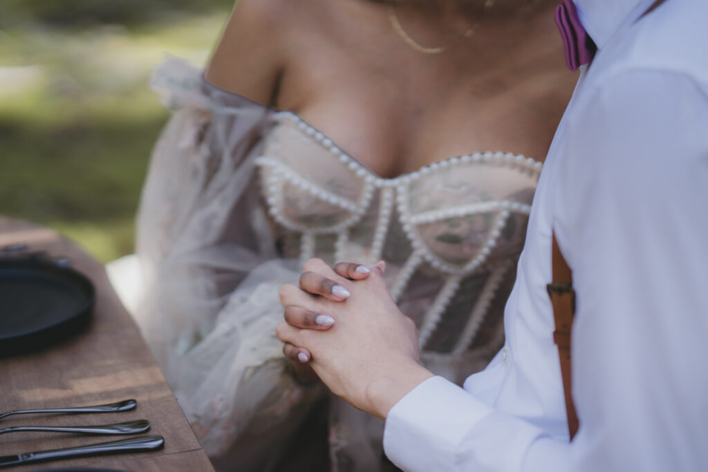 A boho bride and groom holding hands at their table in the forest.