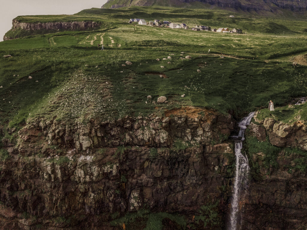A drone shot of a Faroe Islands Elopement couple overlooking a beautiful waterfall at the edge of a cliff. 