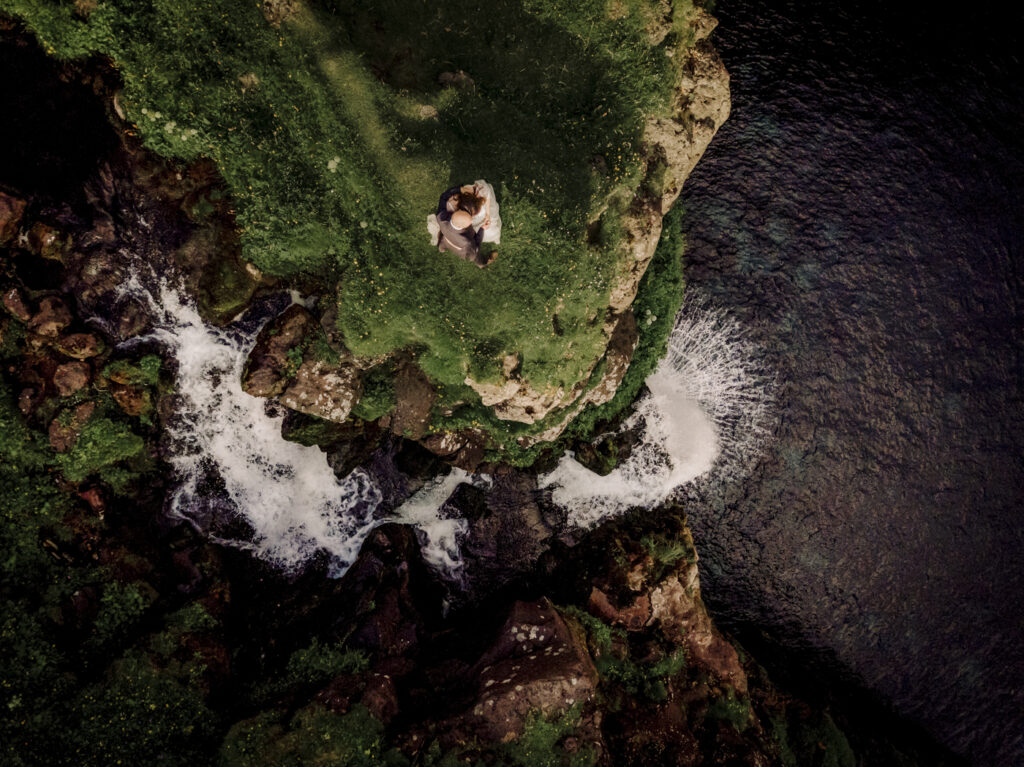 A drone shot of a Faroe Islands wedding couple overlooking a beautiful waterfall at the edge of a cliff. 